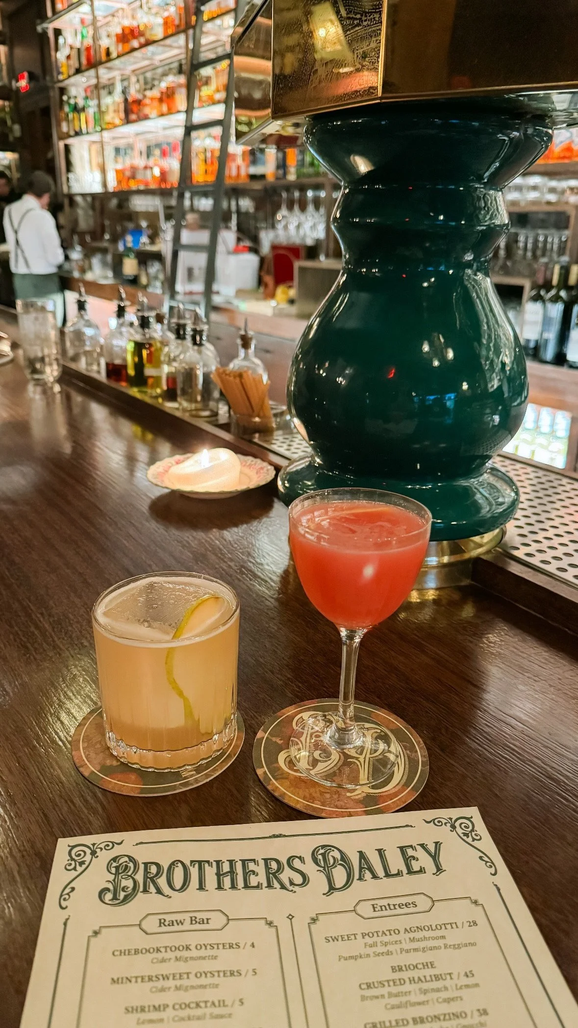 Two cocktails, a yellow lemon drink and a pinkish-red drink, sit on a bar counter with a drink menu called Brothers Daley in the foreground. The bar has a large, dark green sculpture or decorative piece, and various bottles and bar equipment are visible in the background.