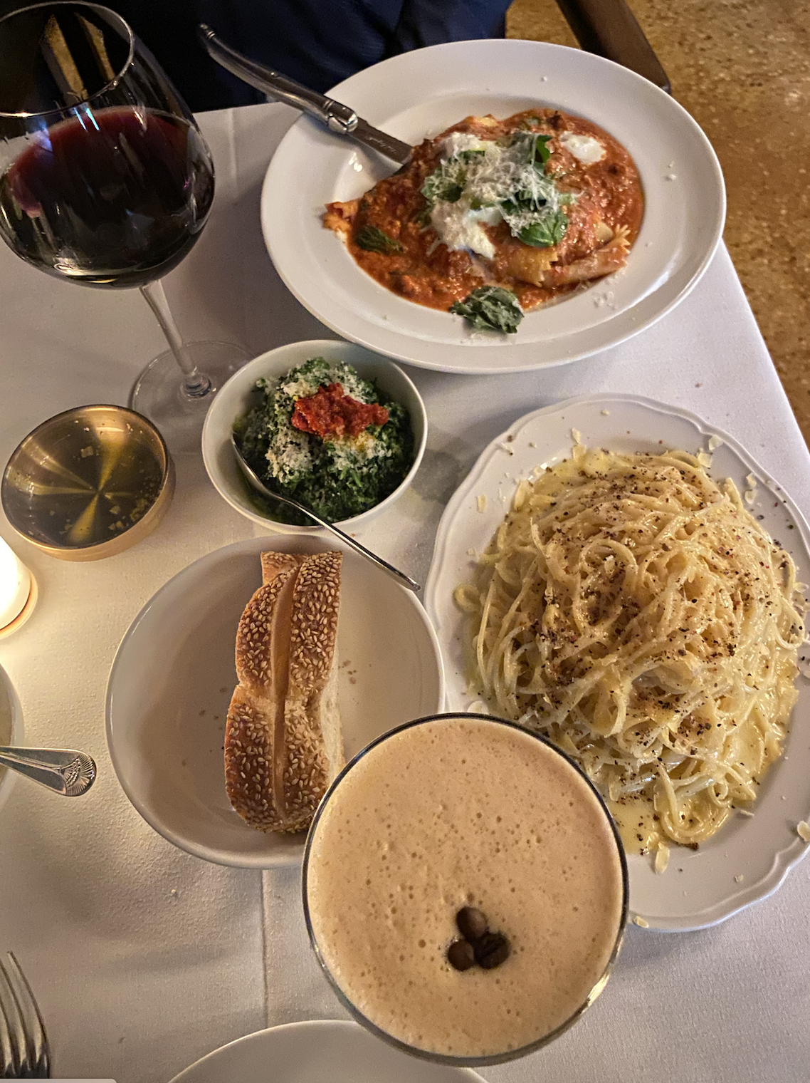 A dinner table with a glass of red wine, a bowl of pasta, a plate of lasagna, a bowl of kale salad, a bread roll, a glass of beer, and salt and pepper shakers.
