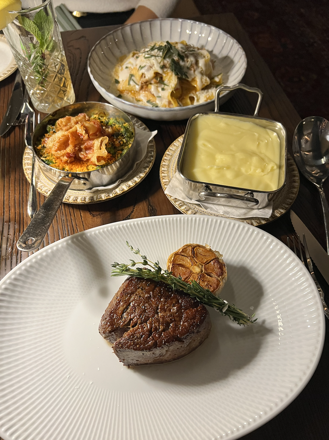 A plated meal featuring a beef steak garnished with a sprig of herbs and a roasted garlic bulb, surrounded by small dishes of mashed potatoes, vegetables, and pasta, with a glass of sparkling water with mint.