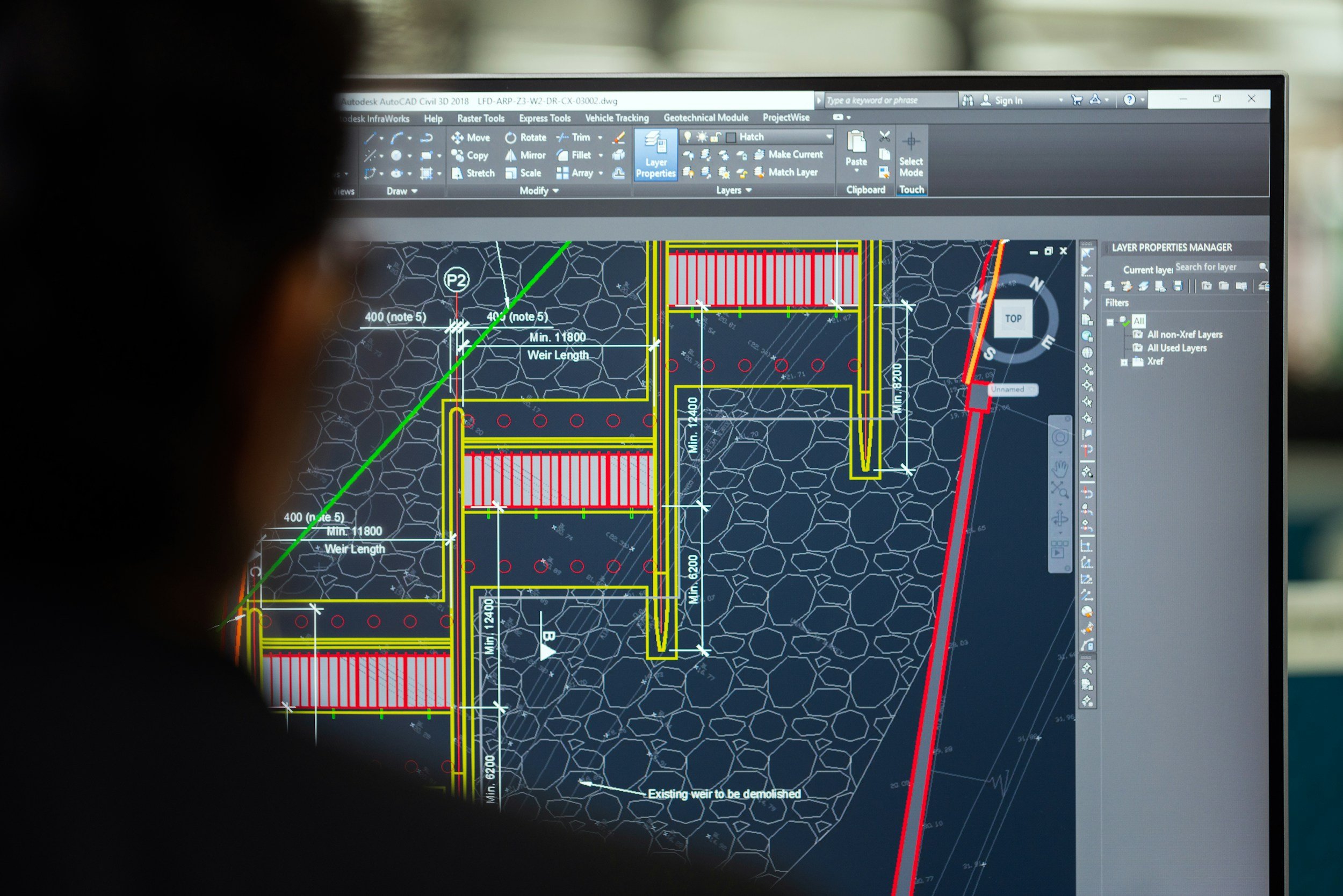 A person looking at a computer screen displaying a detailed civil engineering or construction project plan created with AutoCAD, including measurements, annotations, and technical drawings of a drainage or piping system.