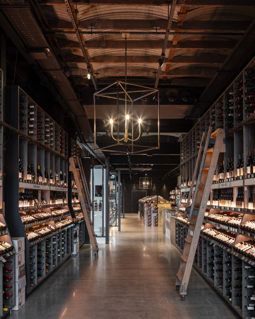 A modern wine shop with shelves of wine bottles, ladders, and contemporary lighting fixtures, including a chandelier and geometric ceiling lights.