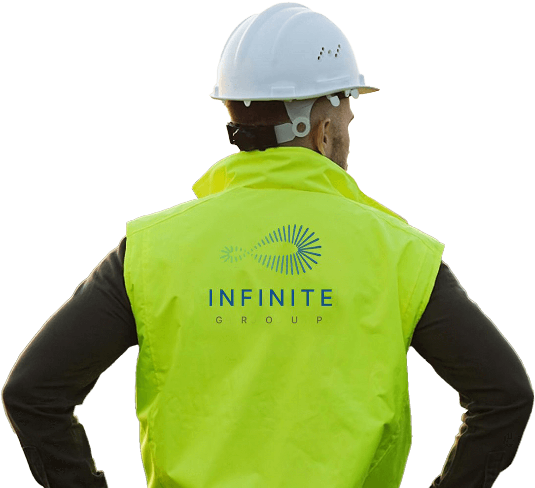 A man wearing a white safety helmet and a neon yellow vest with the 'INFINITE GROUP' logo on the back, standing with his hands on his hips.