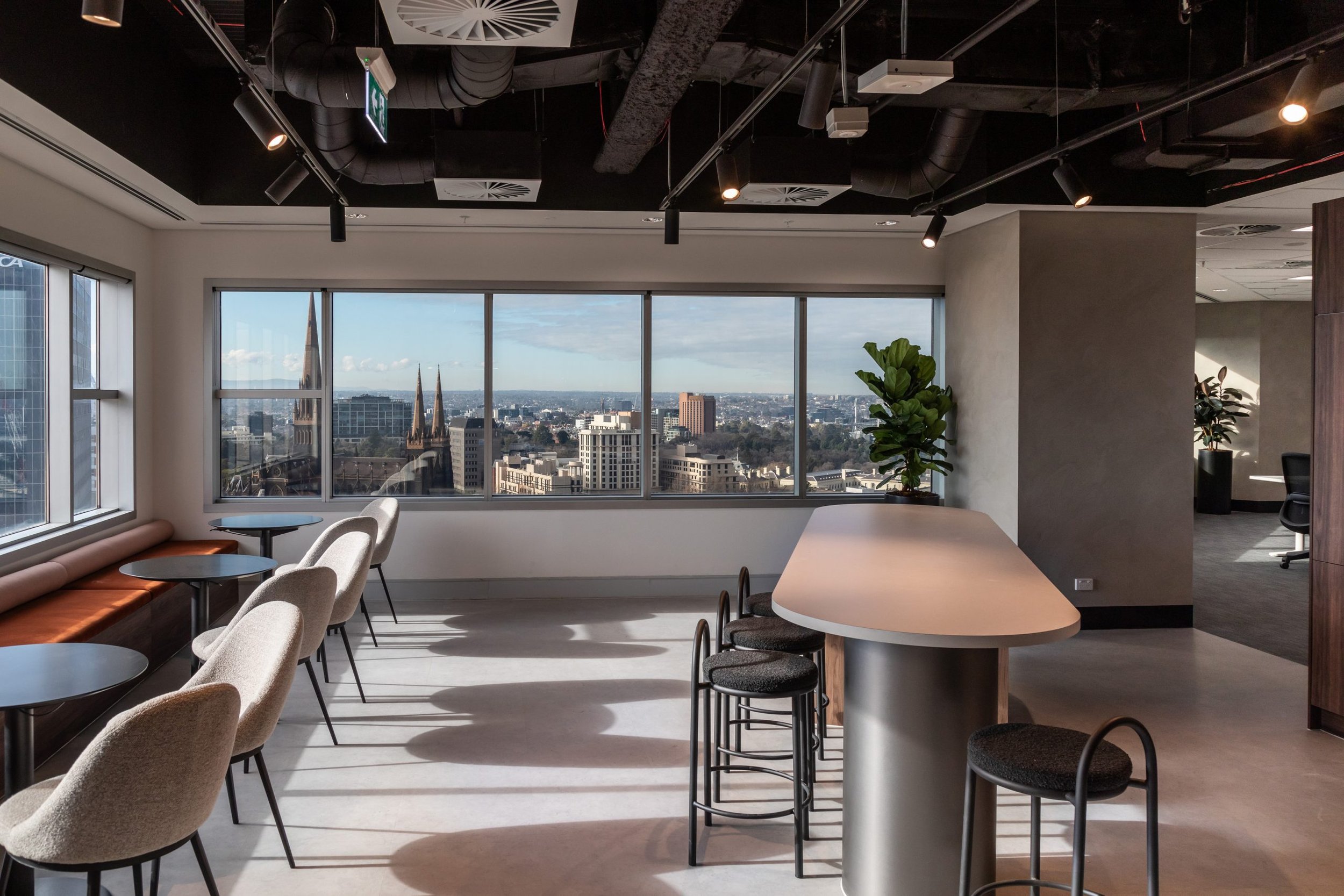 Modern office lounge with large windows, overlooking a cityscape with church steeples, featuring a high table with black bar stools, a row of cozy chairs beside the window, and potted plants, with a black ceiling and track lighting.
