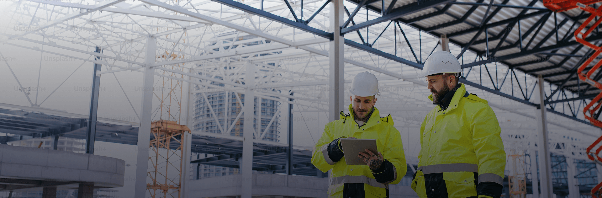 Two construction workers wearing yellow jackets and white hard hats reviewing plans or a tablet on a construction site with a large steel framework and high-rise buildings in the background.