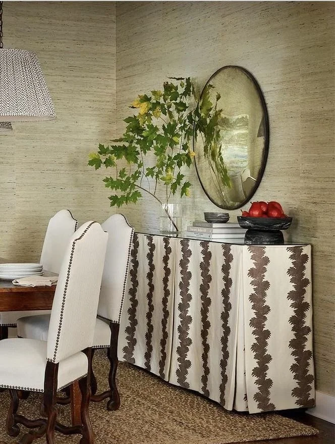 A dining area with a patterned dresser, white upholstered chairs, a mirror, a potted plant, and a bowl of red apples.