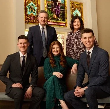 Group of five people, three men and two women, dressed in formal attire, sitting and standing inside a building with stained glass windows in the background.
