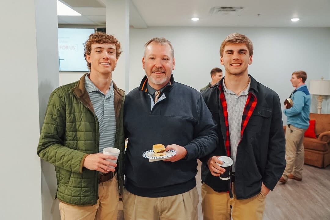 Three men smiling and standing together at a Morningside Valdosta's College and Young Adult Fellowship that meets every other Wednesday evening. Two younger men on either side of an older man, holding coffee cups.