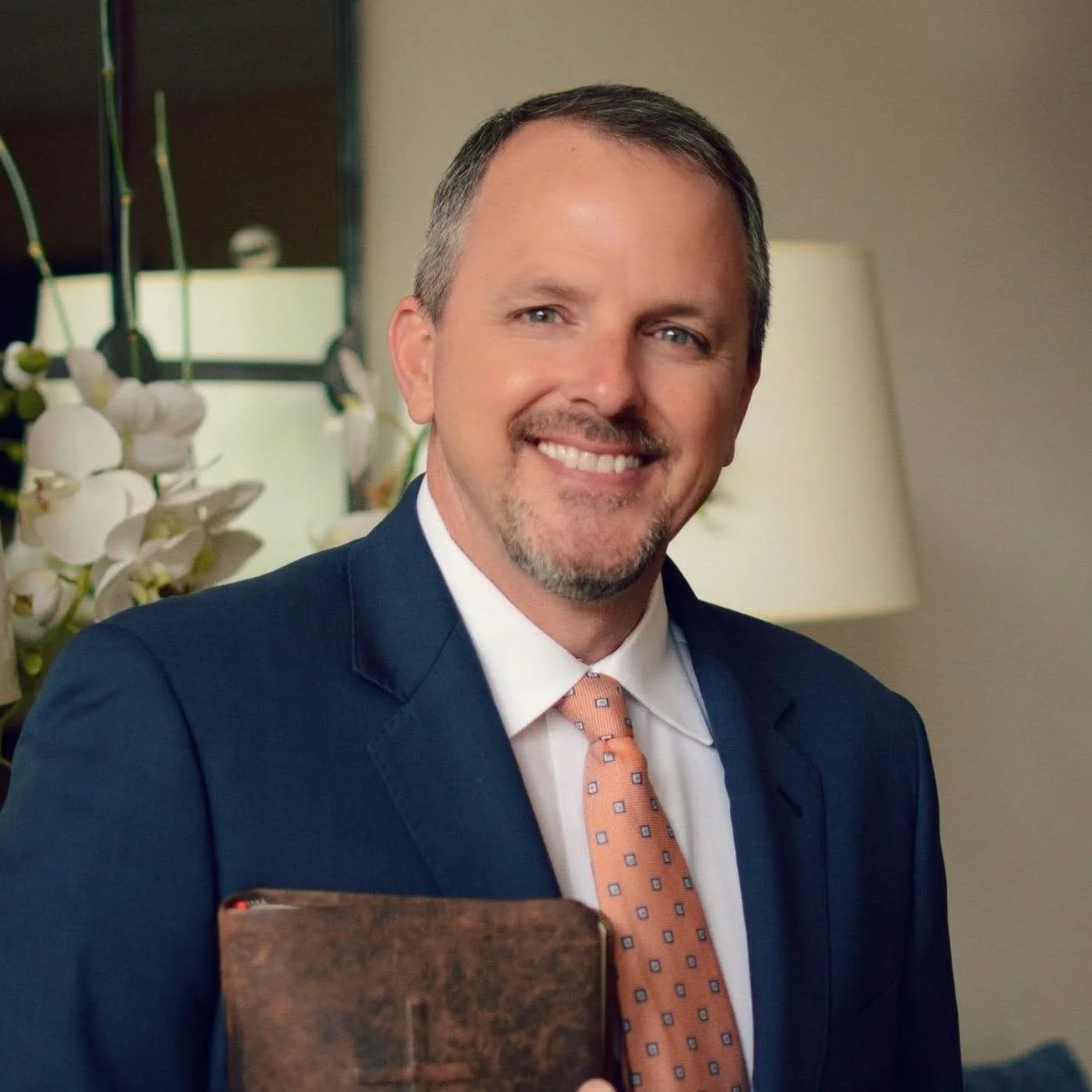 A man smiling, dressed in a blue suit, white shirt, and orange tie, holding a book. Background includes white flowers and a lamp.