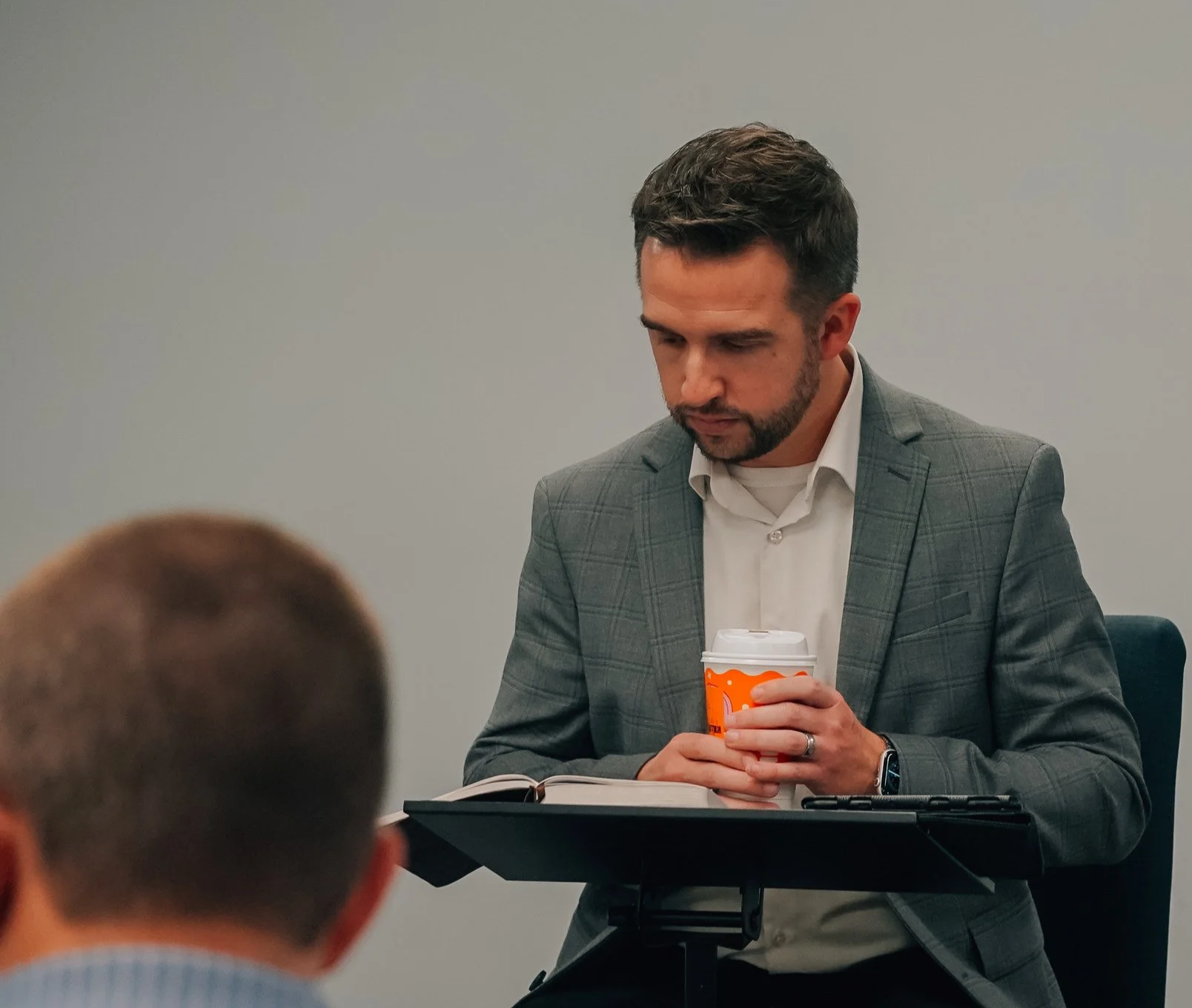 Man in a gray plaid suit holding a coffee cup, standing near a music stand in a meeting room.