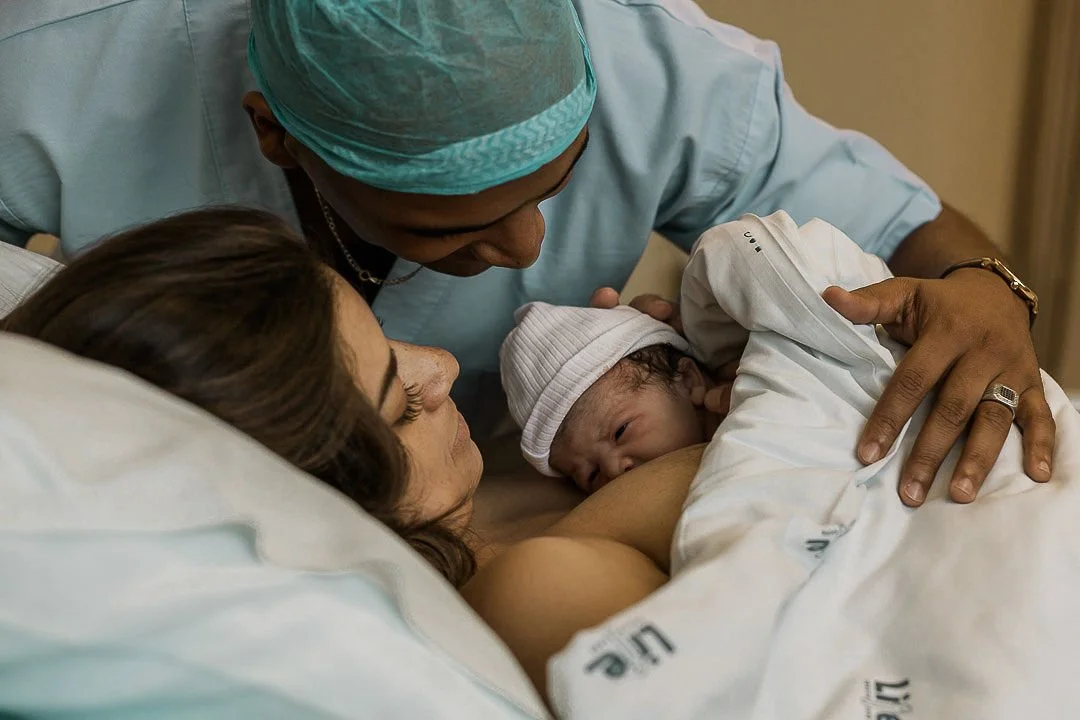 A woman lies in a hospital bed with a newborn baby on her chest, a nurse or doctor is leaning over them, and the baby is wearing a white beanie.