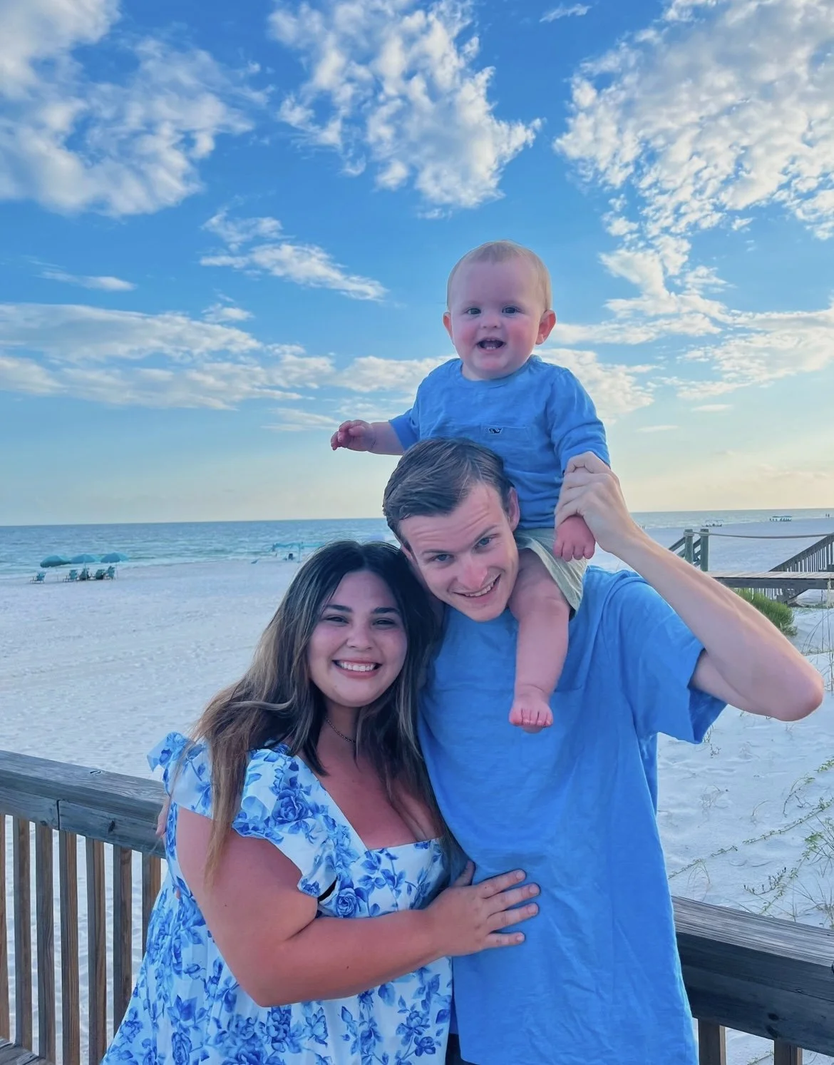 A family of three at the beach: a woman, a man, and a baby. The woman wears a blue floral dress, the man in a blue shirt, and the baby in a blue shirt. The man holds the baby on his shoulders, and all are smiling. The beach with white sand, a railing, and a blue sky with clouds is in the background.