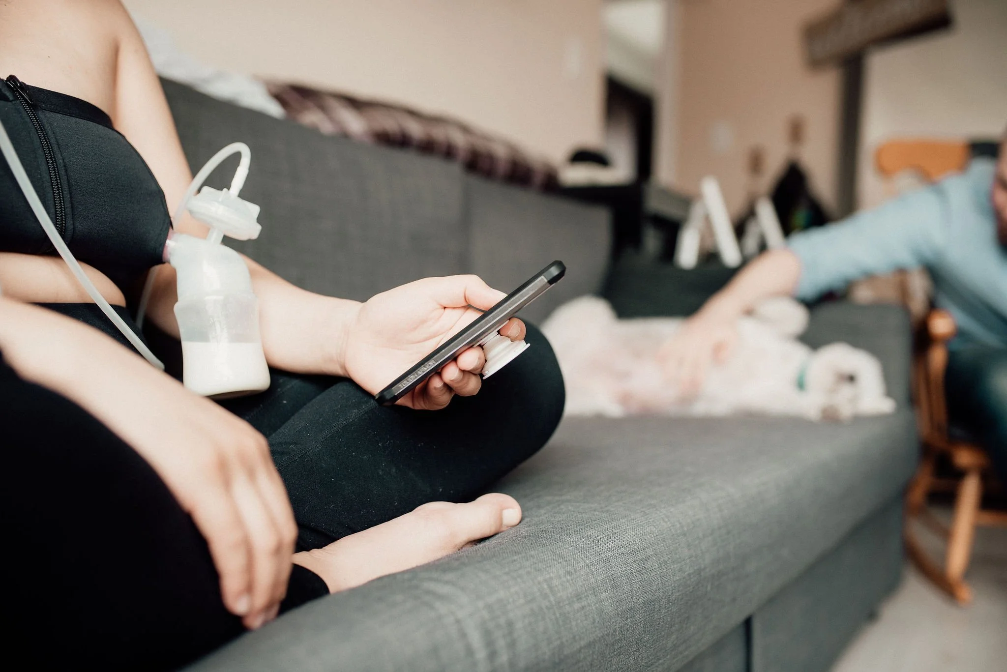 Person sitting on a gray couch holding a smartphone with a nebulizer on their lap. In the background, a person is reaching to pet a small dog lying on the couch.