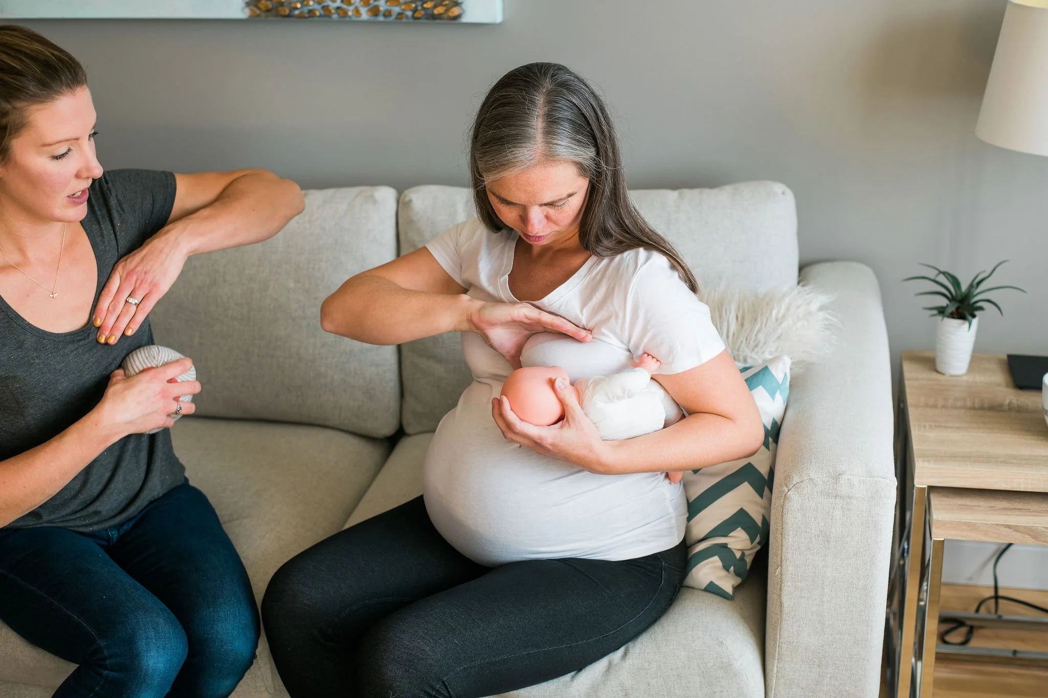 A woman breastfeeding her baby while a friend helps by holding a bottle and offering support in a living room.