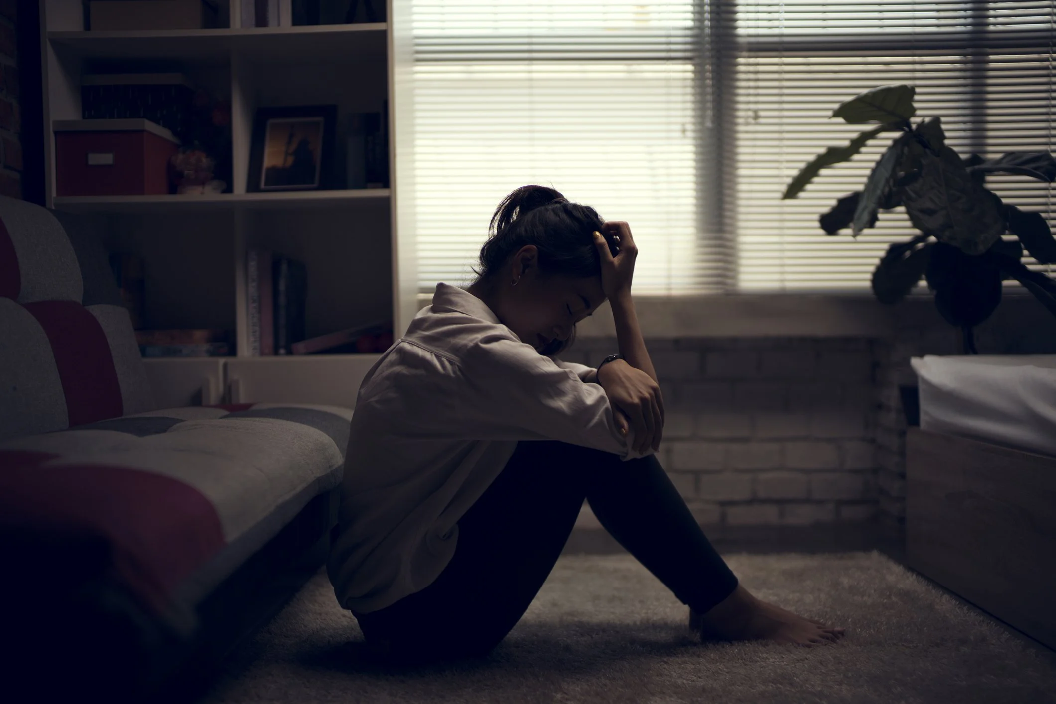 A woman sitting on the floor in a dimly lit room, holding her head in her hand, appearing distressed.