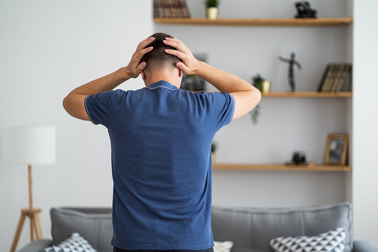 A man standing in a living room with his back to the camera, holding his head with both hands, appearing distressed or stressed.