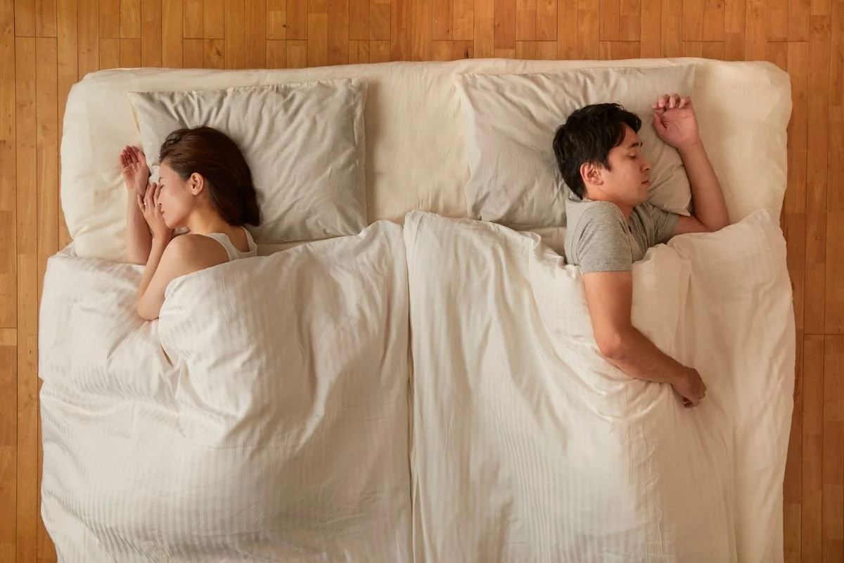 A woman and a man sleeping in beds separated by a small gap, viewed from above. The woman has long brown hair and is lying on her side, covering her face, with a pillow behind her. The man has short dark hair, is lying on his side, and appears to be sleeping peacefully.