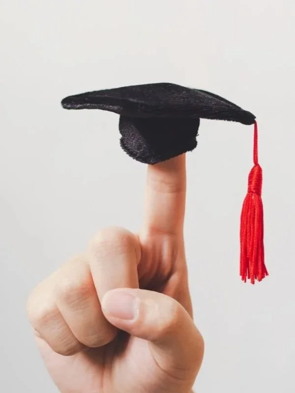 A person's hand balancing a small black graduation cap with a red tassel on their index finger.