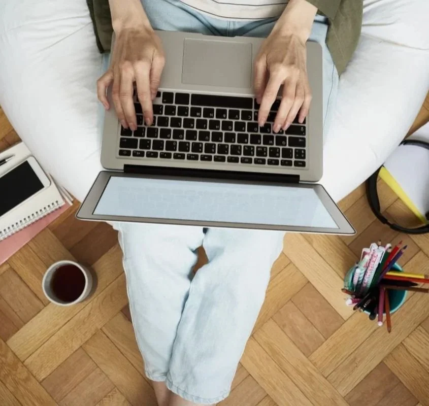 Person sitting on a bed with their legs crossed, working on a laptop. Nearby, there is a cup of coffee, a notebook, and a container filled with various colored pens.