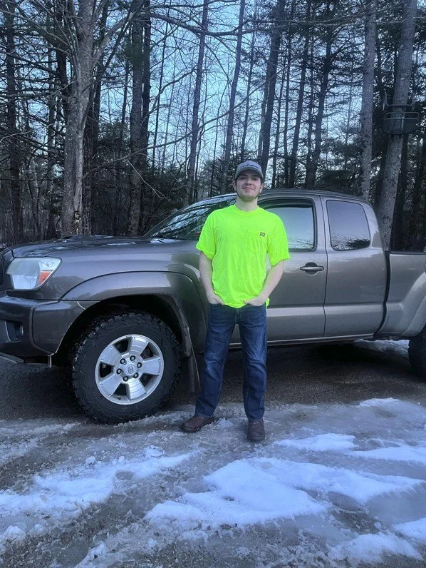 A young man in a bright yellow-green shirt and jeans standing next to a gray SUV in a wooded area with some snow on the ground.