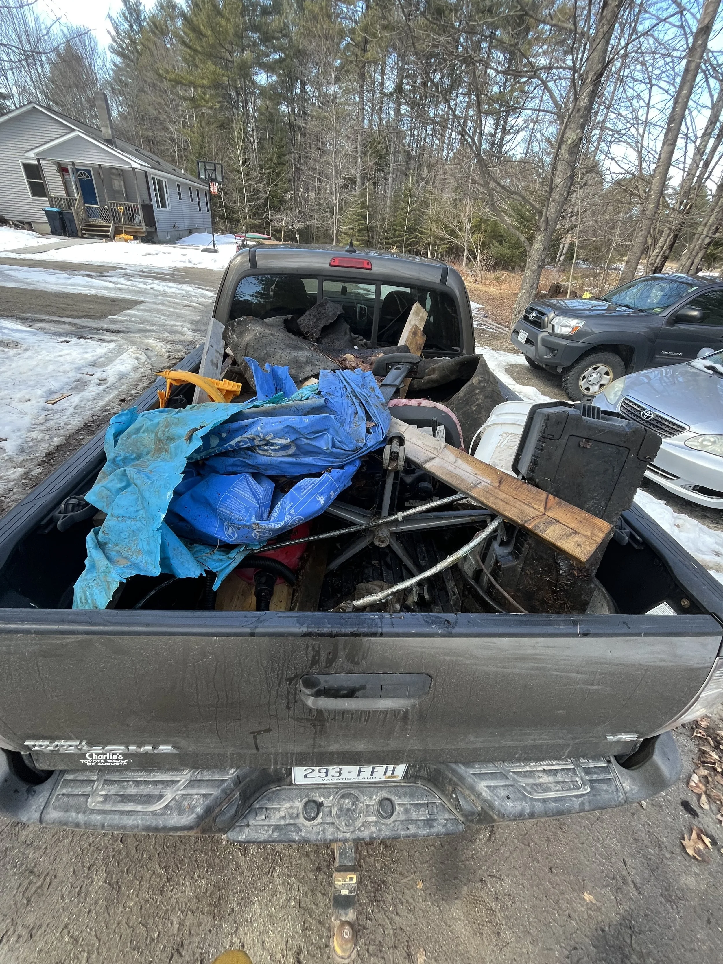 Back of a pickup truck filled with various items, including blue tarps, wood, metal rods, and plastic containers, parked in a driveway with snow and other cars nearby.