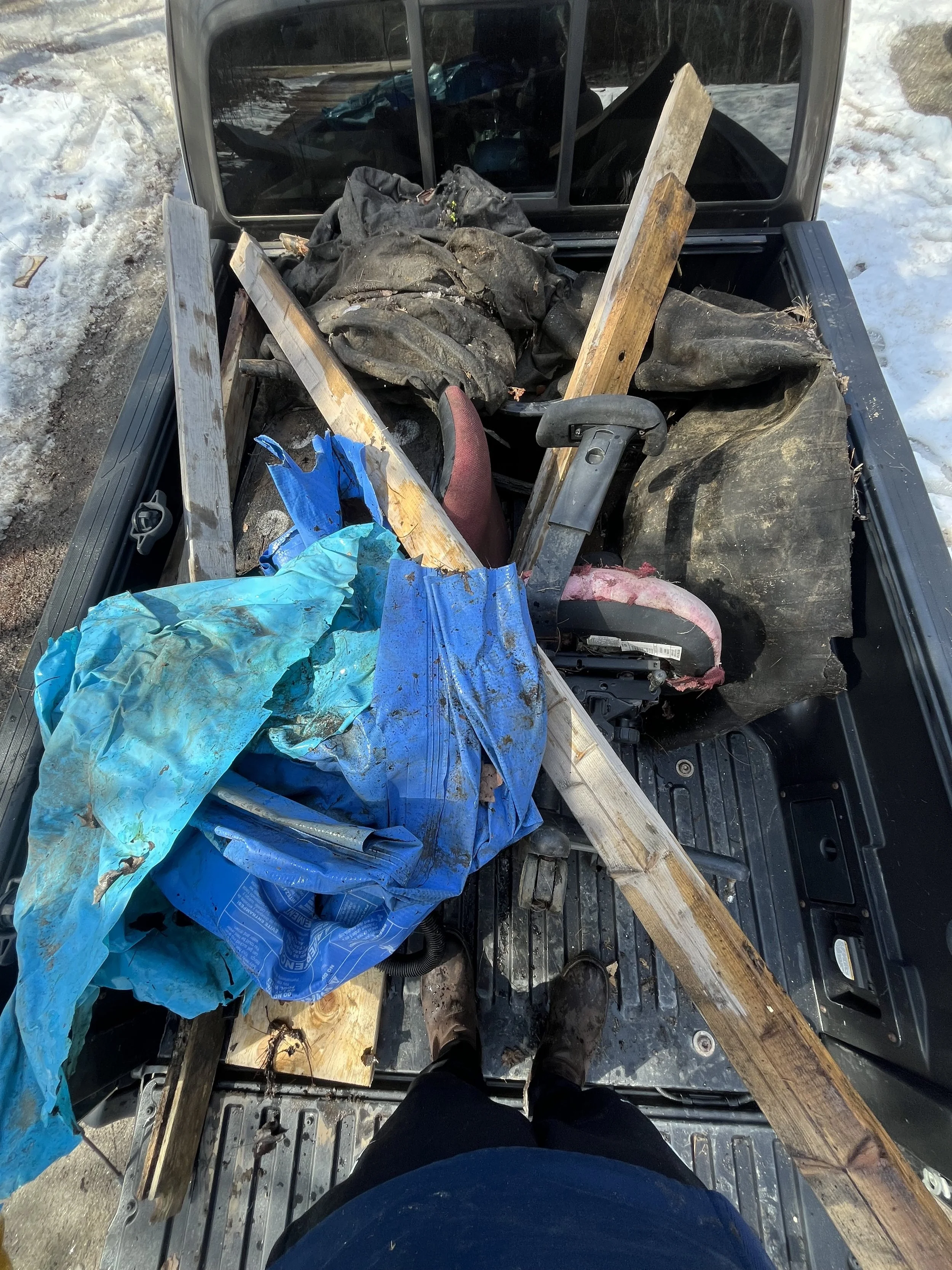 View from above of a pickup truck bed filled with wood, a blue tarp, and various tools and materials, with person standing at the edge wearing dark pants and boots, surrounded by snow.