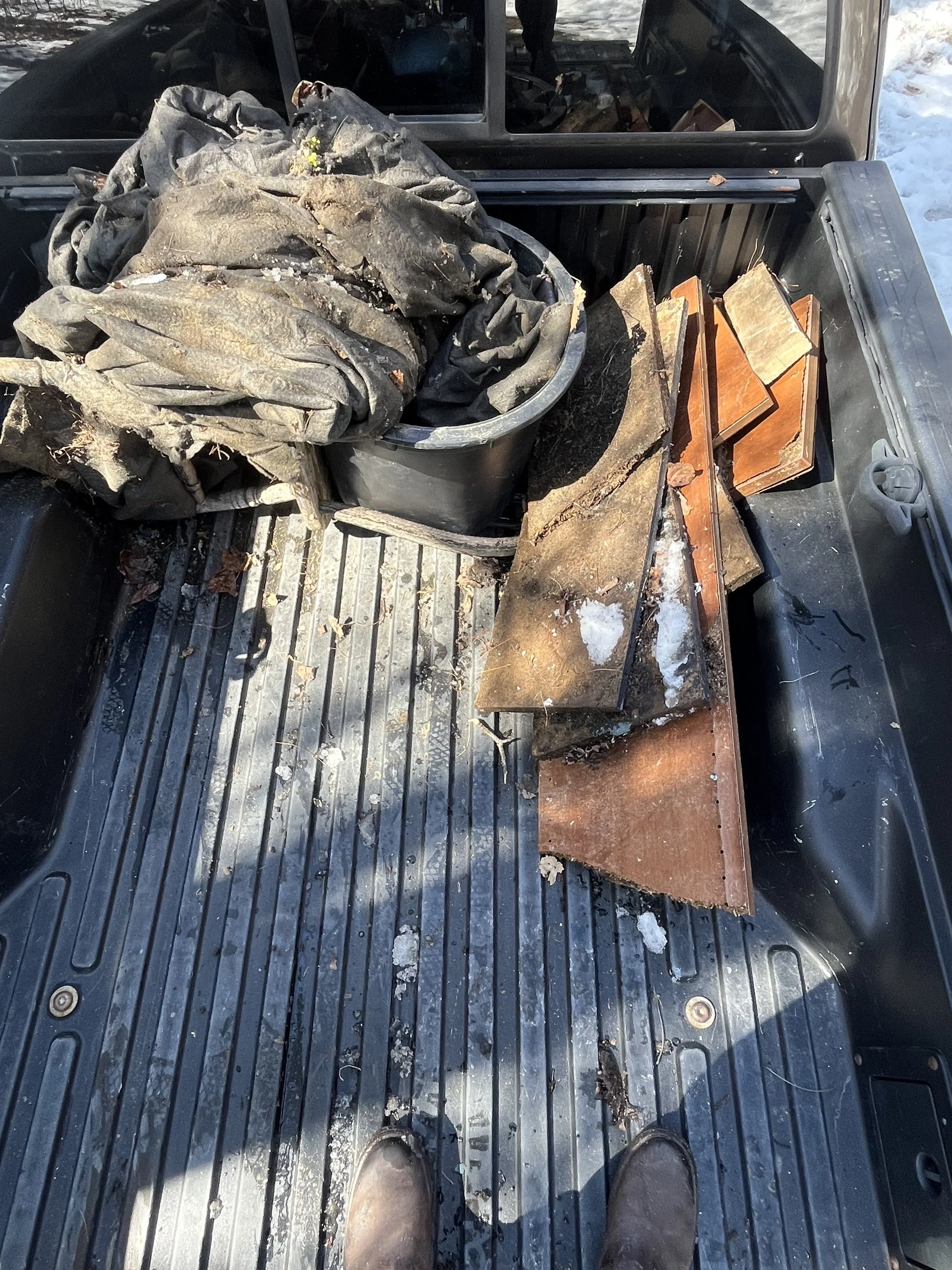 Pickup truck bed filled with a dirty tarp, a black container, rusted metal sheets, and wooden planks, with snow outside.