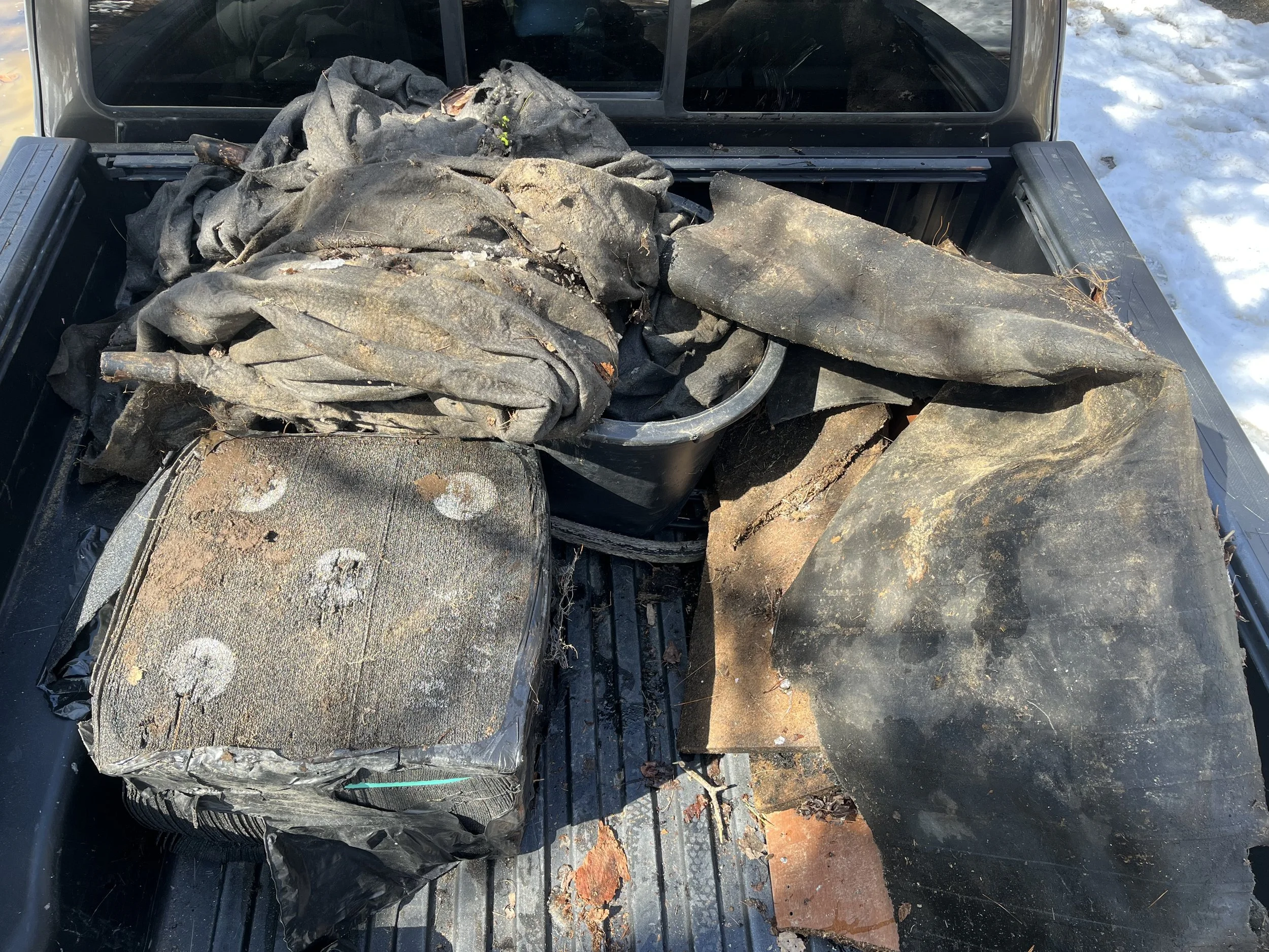Truck bed filled with old, dirty, and torn black vehicle covers or tarps, some folded and some partially unfolded, with dirt, debris, and signs of wear. Snow is visible on the ground around the truck.