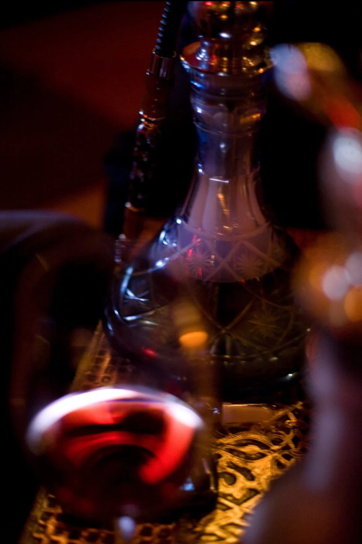 Close-up of a glass hookah with intricate designs on its base, surrounded by colorful blurred lights, and a small glass of red liquid in front.