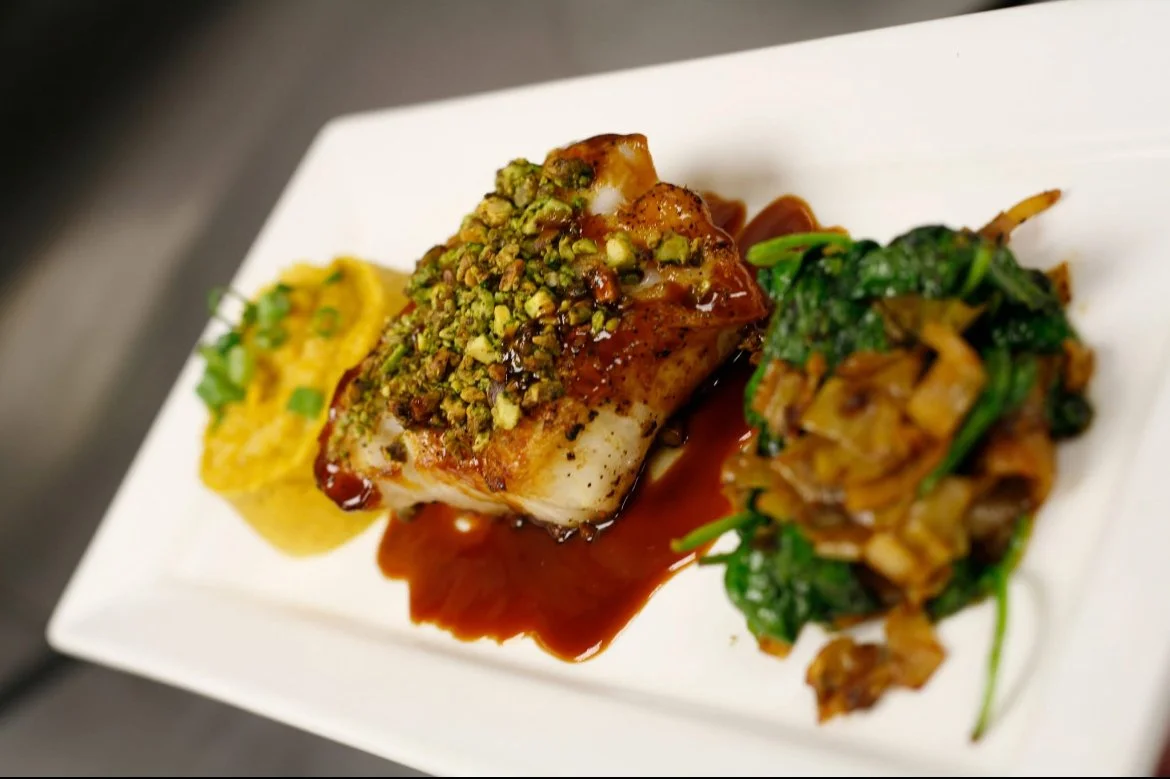 A plated dish with a piece of fish topped with chopped pistachios, served with a green vegetable side and a yellow side, all on a white rectangular plate.