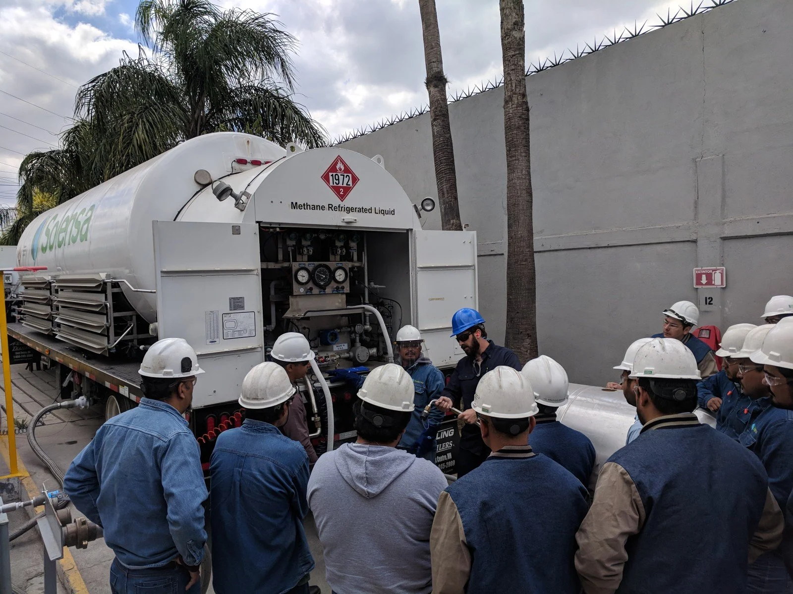 Group of workers in safety helmets around a methane refrigerant liquid tank truck during a safety briefing or demonstration outdoors. The truck is labeled with hazard signs and the company name Soersa.