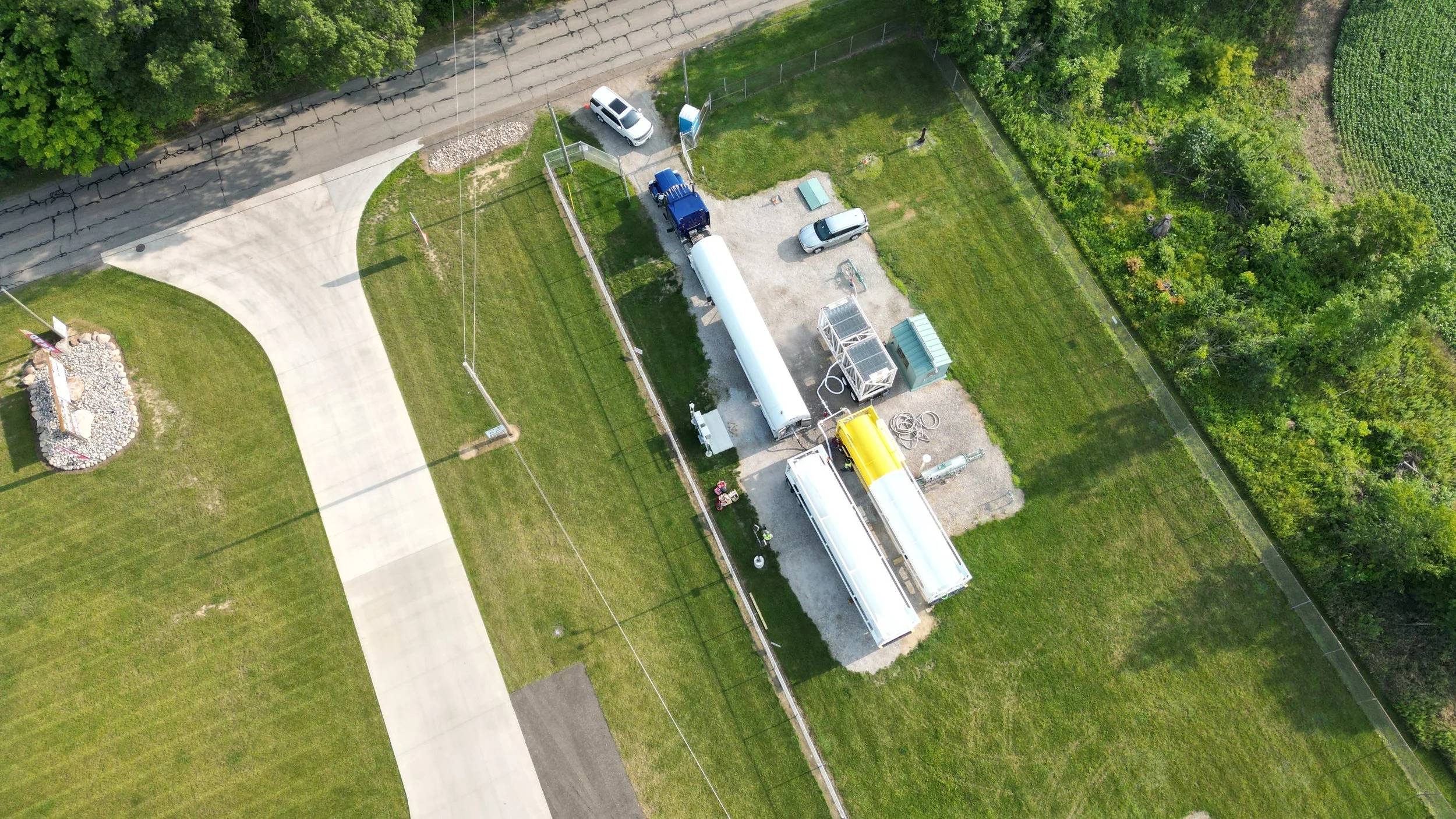 An aerial view of a small private property with a driveway, a parking area, and a grassy yard surrounded by trees and some agricultural land in the background. There are several vehicles parked on the driveway and yard, including a large white tank and other construction or utility equipment.