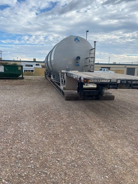 Large cylindrical tank on a flatbed trailer in an outdoor lot, with gravel ground, some buildings, a green dumpster, and a cloudy sky in the background.