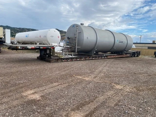 Two large cylindrical tanks on flatbed trailers parked on gravel ground with a partly cloudy sky and distant mountains in the background.
