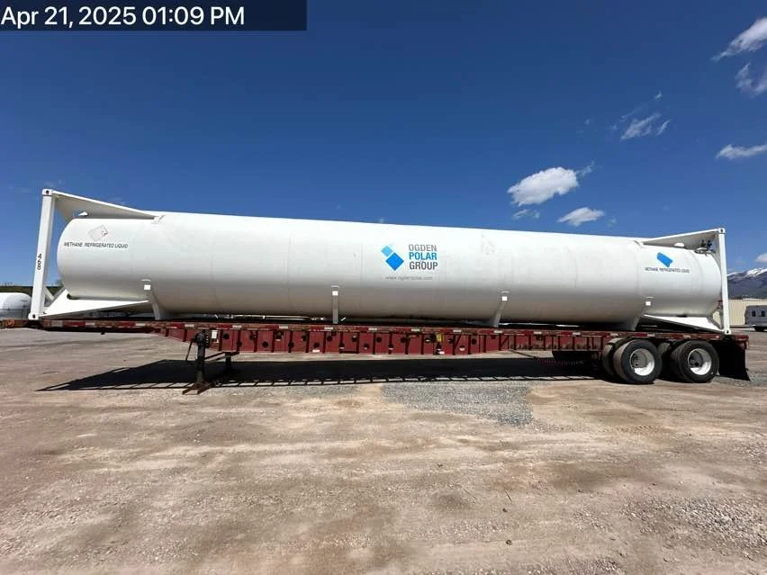 A large white ethanol storage tank with Ogden Polar Group logo on a red flatbed trailer, parked on a dirt lot under a partly cloudy blue sky. Mountains are visible in the distance.