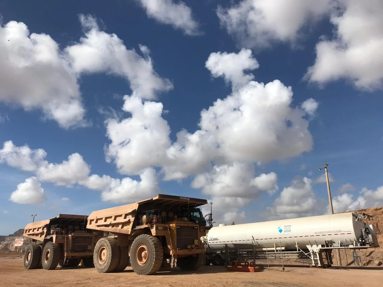 A large dump truck parked next to a white tanker trailer at a mining or construction site under a partly cloudy blue sky.
