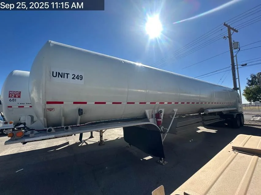 A large white tanker trailer with 'UNIT 249' labeled on its side, parked outdoors under a bright sun with a clear blue sky, power lines, and a few trees in the background.