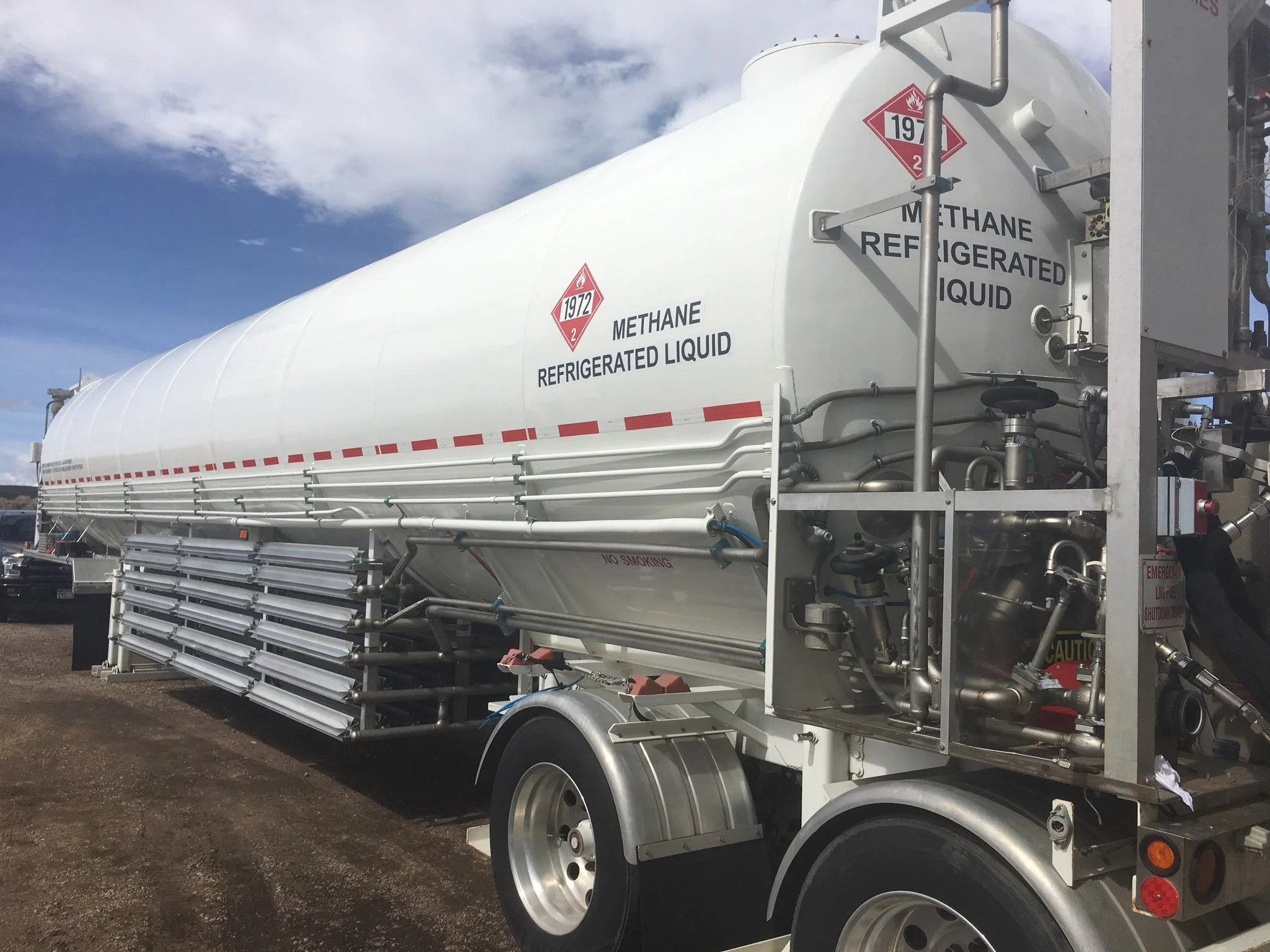 A large white tanker truck containing liquid methane, labeled with hazard symbols and warning signs, parked outdoors under a partly cloudy sky.