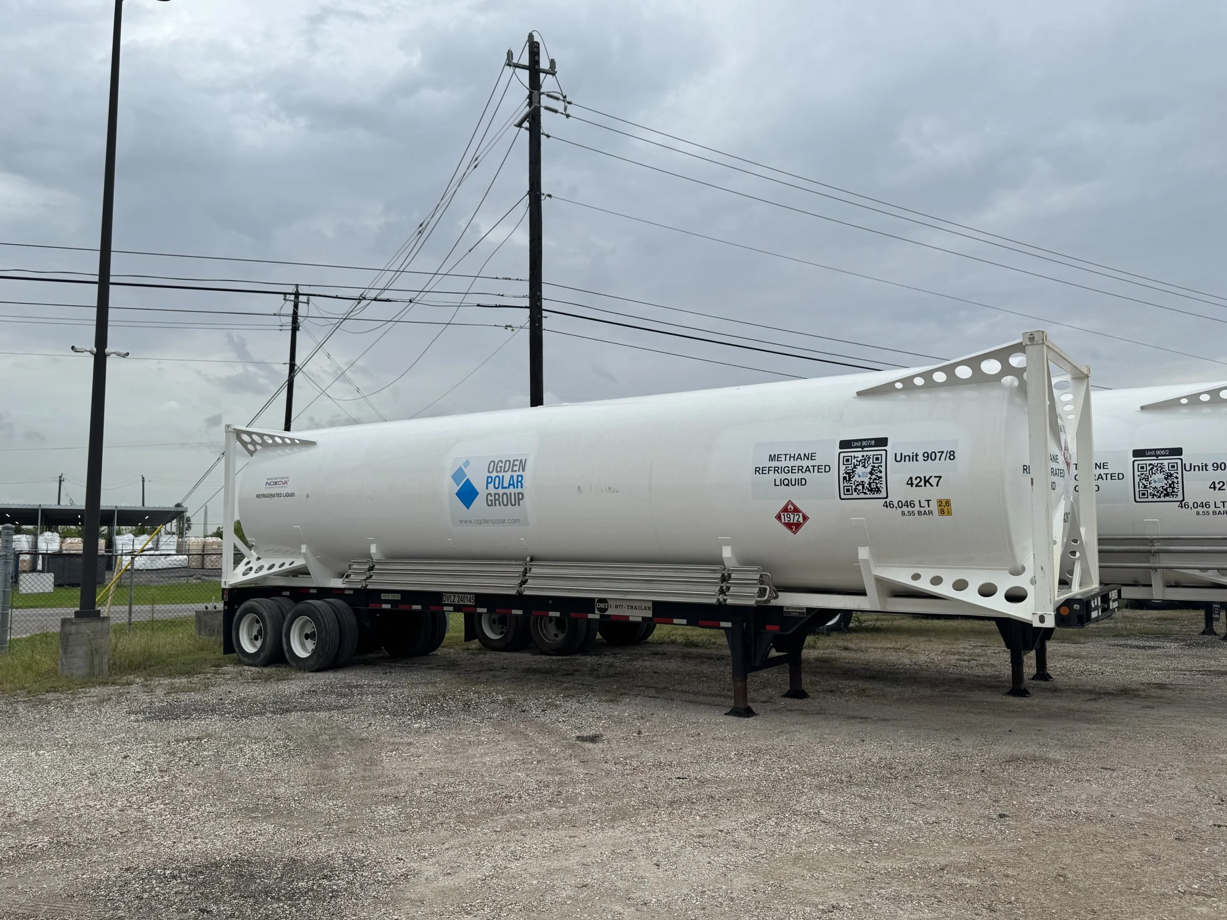 A white tanker trailer with Ogden Polar Group logo, labeled as containing refrigerated liquid methane, parked outdoors on a gravel lot with utility poles and a fence in the background under an overcast sky.