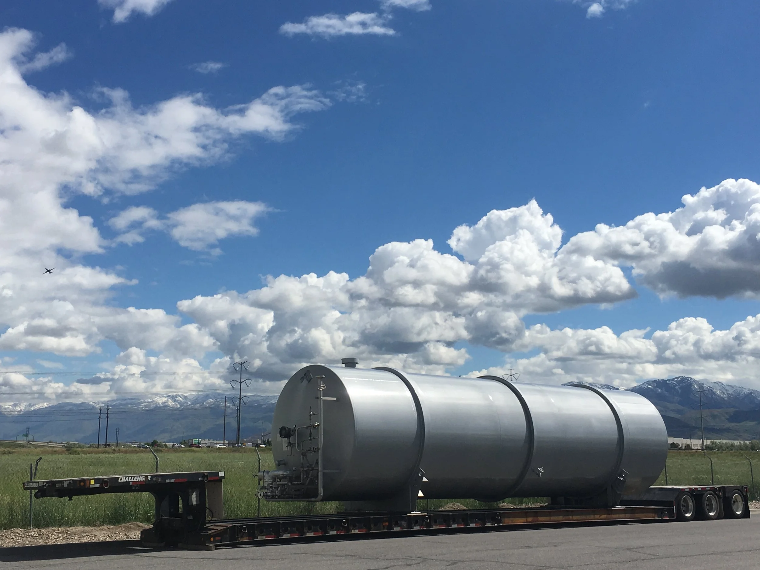 Large silver cylindrical tank mounted on a flatbed trailer on the side of the road, with mountains, power lines, and a blue sky with white clouds in the background.