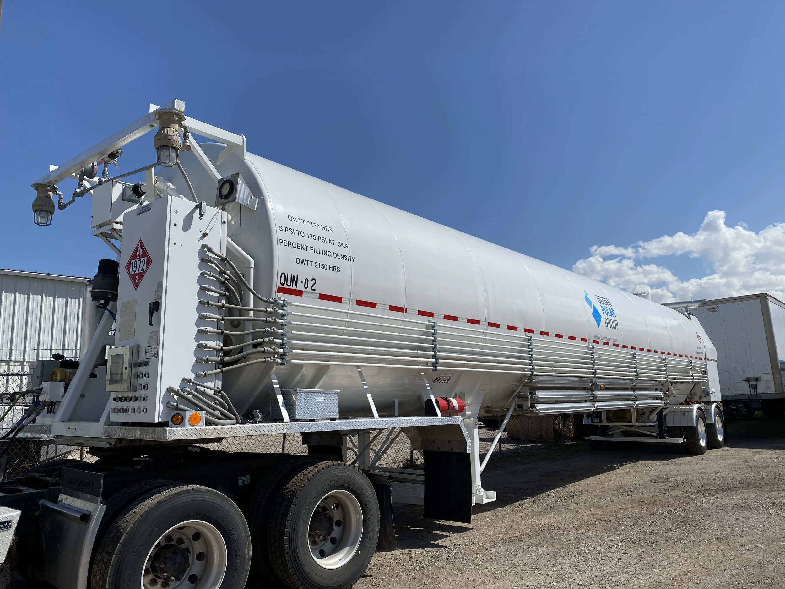A large white tanker trailer parked outdoors under a clear blue sky with a few clouds, used for transporting liquid fuels, with various tire and safety markings.