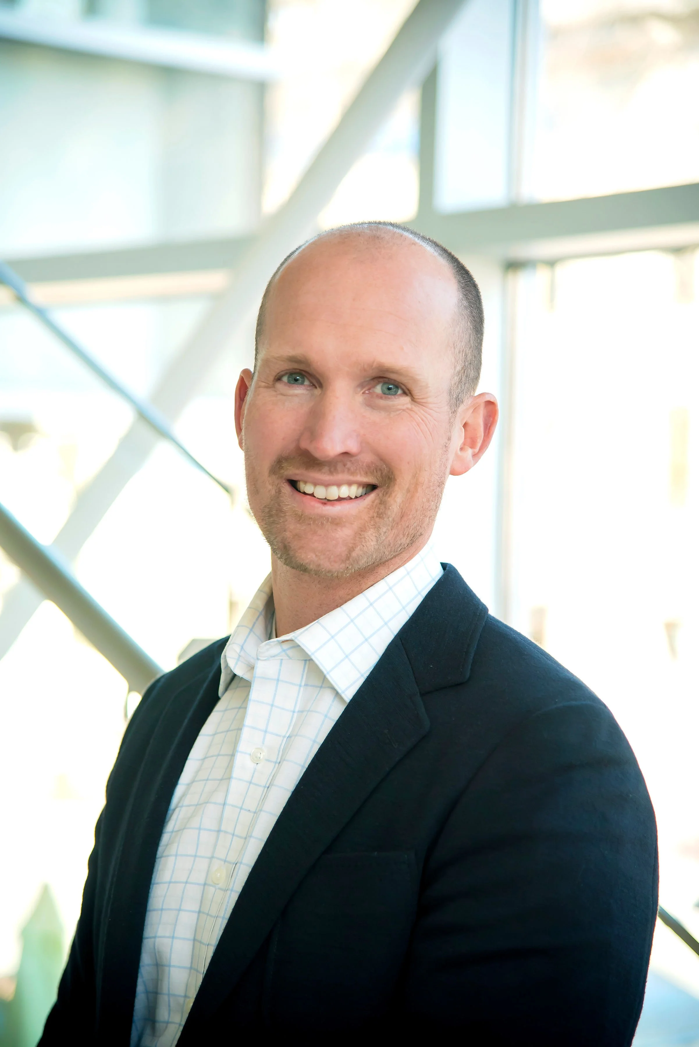 A man with a receding hairline and a light beard, smiling, wearing a dark blazer over a white shirt with a light checkered pattern. He is standing indoors near large windows with natural light.