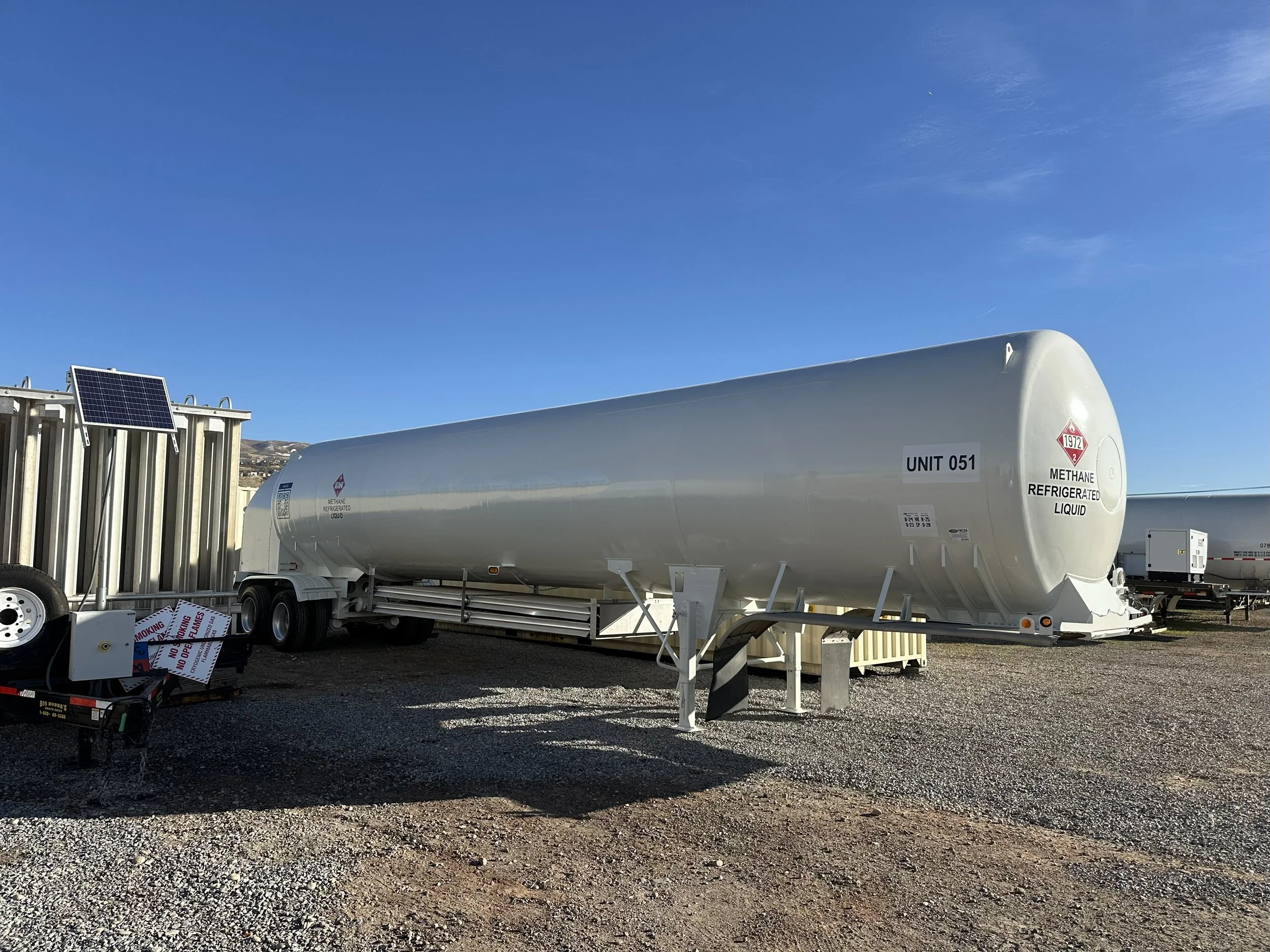 A large white methane refrigerated liquid storage tank truck with a label near the front that reads 'Unit 051' is parked on a gravel lot under a clear blue sky. There is a small solar panel mounted on a nearby structure and additional tanks in the background.
