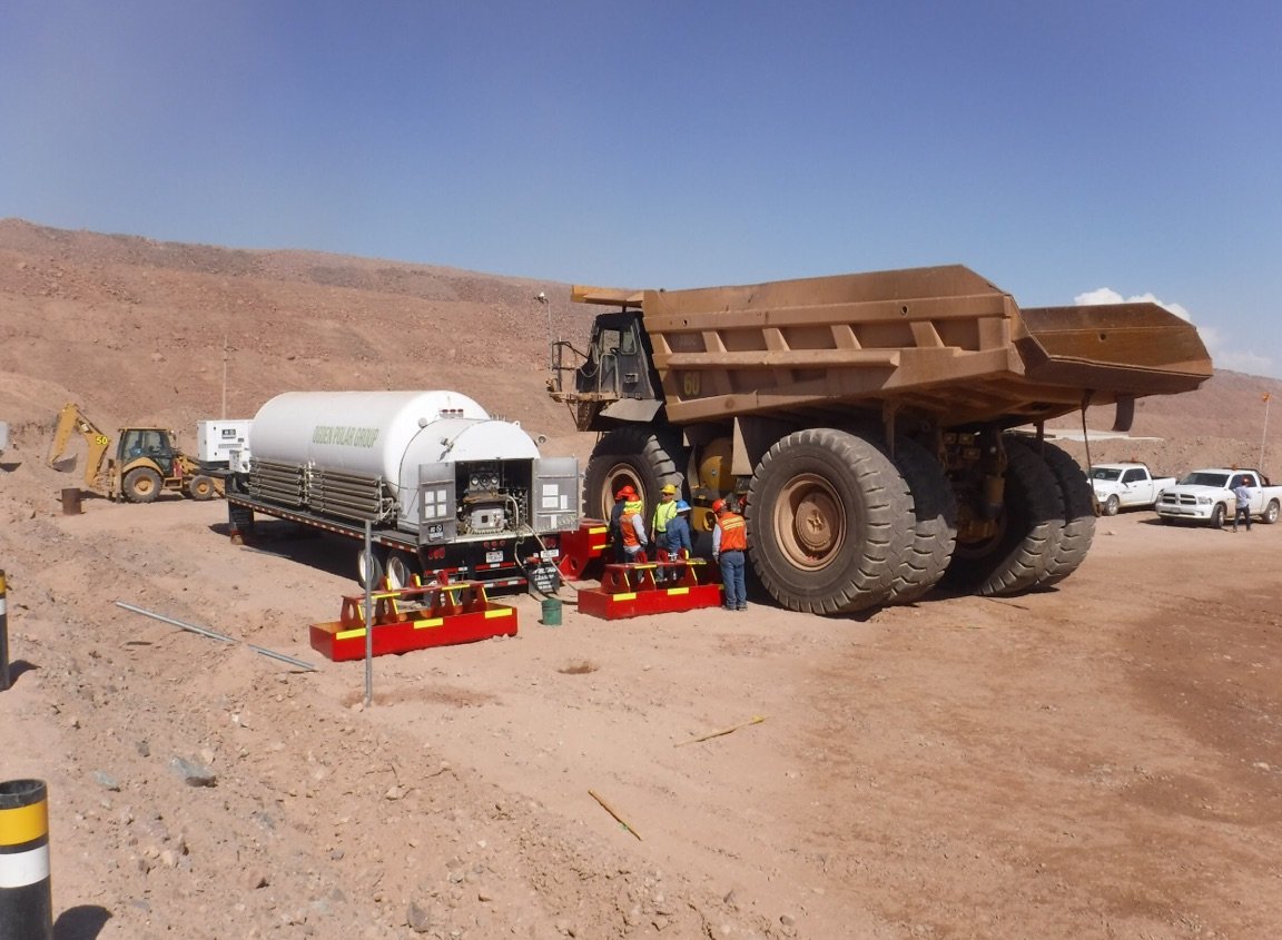 Construction workers operating heavy machinery at a construction site in a desert landscape, with a large dump truck, a water tanker, and a backhoe in the background.