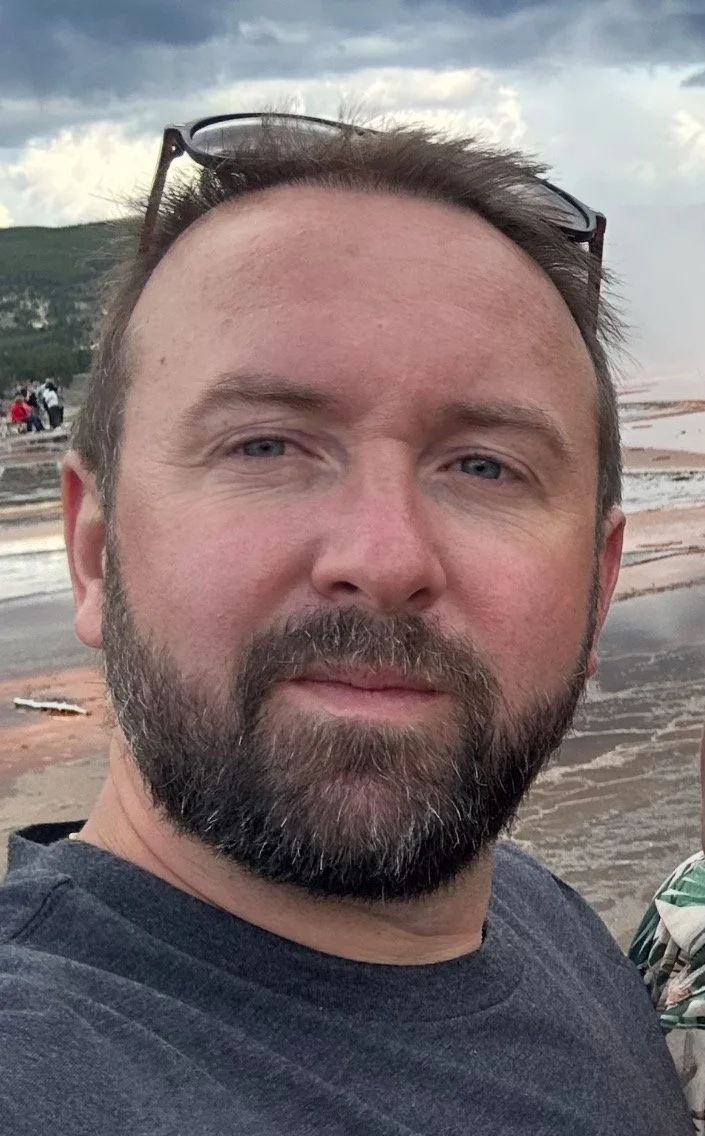 A man with a beard and glasses resting on his head, taking a selfie outdoors with a cloudy sky and water in the background.