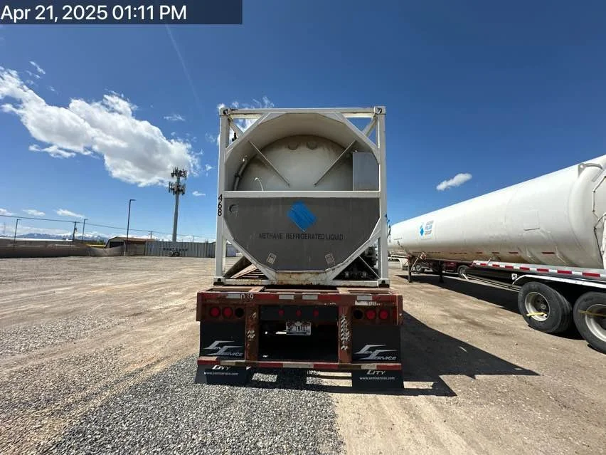 Rear view of a semi-truck trailer carrying a cylindrical container labeled "Methane Refrigerated Liquid" parked in an open lot with other trucks, under a partly cloudy sky.