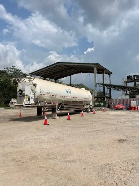 Large white tanker truck parked under a metal canopy with orange traffic cones around it, in an open area with gravel ground and some trees and industrial structures in the background.