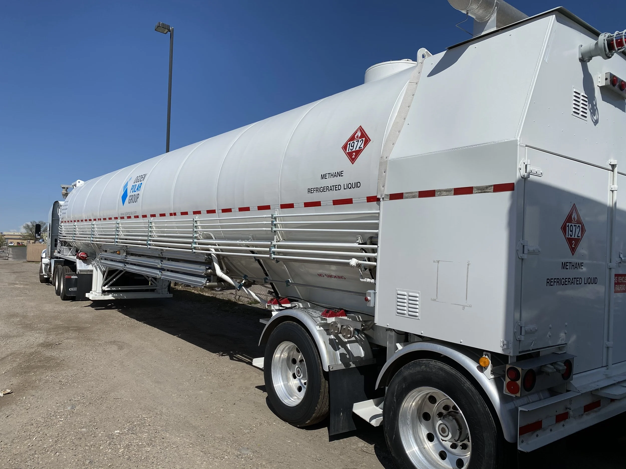 A large white tank trailer labeled with hazardous material signs and the words "Methane Refrigerated Liquid," parked on a dirt lot under a clear blue sky.