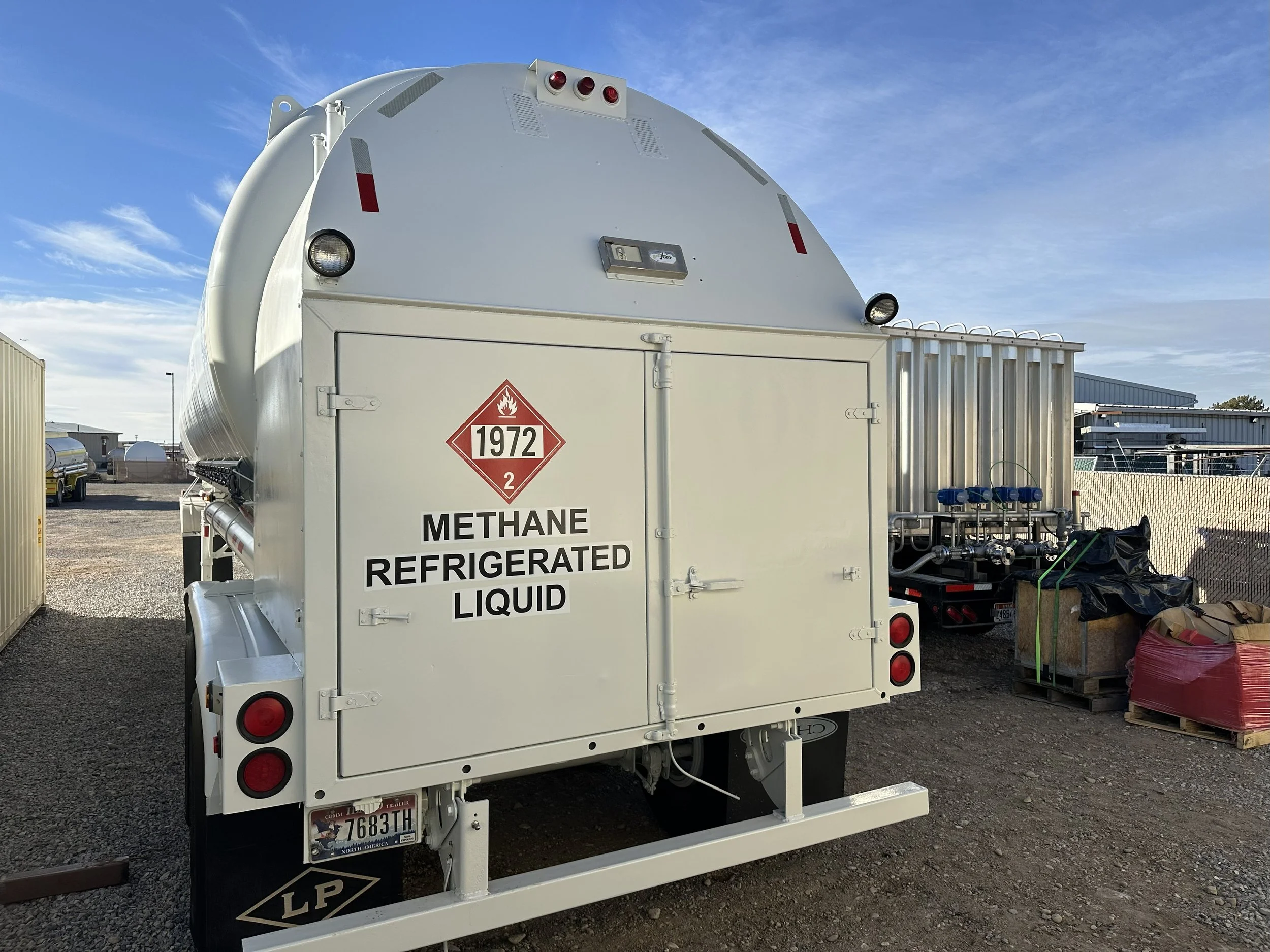 A white tanker truck with a warning label indicating it contains refrigerated liquid methane, parked outdoors on a gravel lot under a blue sky.