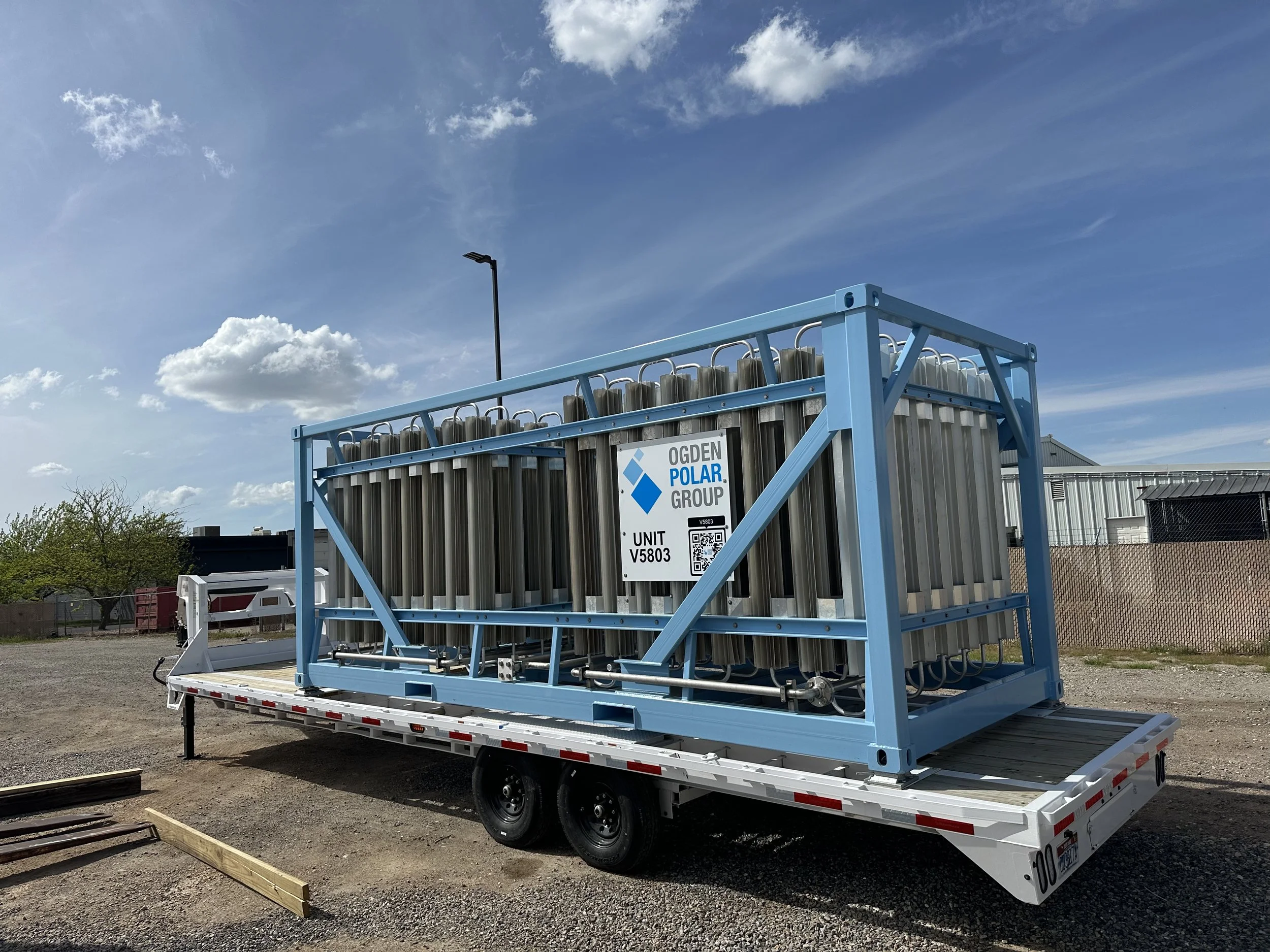 A large industrial machine with multiple vertical metal cylinders mounted inside a blue metal frame on a flatbed trailer. The trailer is parked outdoors on a dirt surface under a partly cloudy sky. There is a sign on the machine reading 'OGDEN POLAR GROUP' and a QR code.