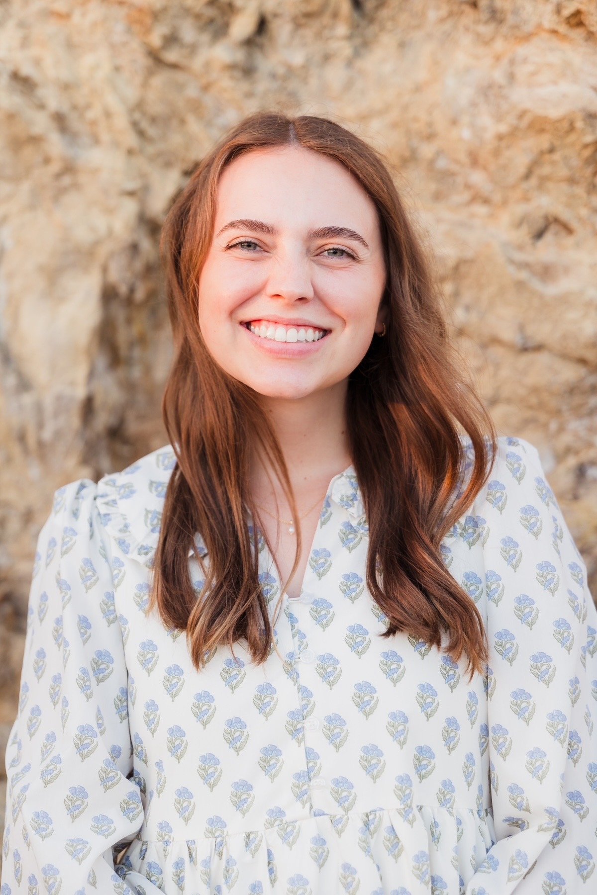 Portrait of a smiling woman with long wavy brown hair, wearing a white blouse with a blue floral pattern, standing outdoors with a rocky background.